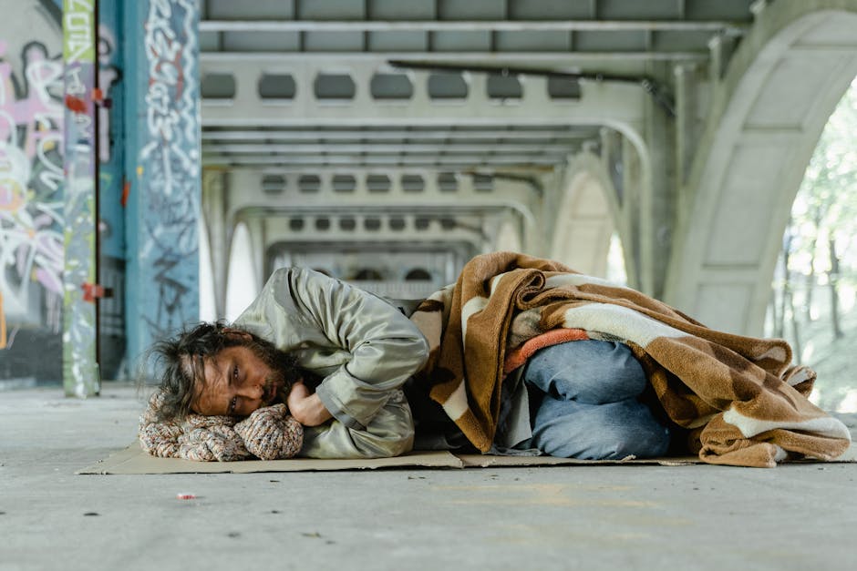 A homeless man lies under an urban bridge, wrapped in a blanket, symbolizing poverty and survival.